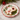 A scalloped ceramic holiday plate with a red rim and holly and berry print, styled with two Christmas cookies on a light marble surface with evergreen branches in the background.
