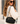 A woman wearing a black crossbody bag with a white and black striped strap and gold hardware, photographed outdoors against a blurred architectural background.