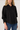 A woman wearing a black long-sleeve button-up blouse with gathered cuffs and blue jeans, photographed against a plain light gray studio background.