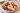 A large white ceramic serving platter with a red scalloped rim and a Mediterranean-style charcuterie board design, filled with cucumber slices, cherry tomatoes, olives, salami, cheese cubes, crackers, and a small bowl of white dip on a marble table.