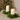 Two cream-colored LED pillar candles placed in a green evergreen garland on a light beige table, with a blurred Christmas tree in the background.