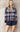 A woman modeling a long-sleeve button-up blouse with a navy blue plaid pattern and a red, white, and blue striped V-neck placket, paired with navy shorts, standing against a textured concrete wall.