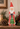 A Santa Claus figurine wearing a red coat and hat with a white beard, holding a green Christmas tree, displayed on a wooden surface with pinecones, evergreen branches, and a lit candle in the background.