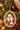 An oval Christmas ornament featuring a painted portrait of Santa Claus in a red suit and pink glasses, hanging from a gold-toned frame on a decorated Christmas tree with lights and ornaments.