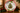 A scalloped ceramic Christmas plate displayed on a gold stand on a wooden table, surrounded by holiday greenery, red ornaments, and lit candles.