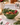 A green ceramic bowl with a leaf-like scalloped rim, filled with mixed nuts and cranberries, set on a marble countertop in a decorated holiday kitchen scene.