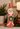 A distressed red Santa Claus figurine holding a green wreath, styled on a tabletop with a lit candle, potted greenery, and red berries in the background.