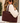 A woman wearing dark sunglasses and a cream ribbed turtleneck sweater carries a large dark burgundy suede tote bag with gold hardware and the brand name 'KATIE LOXTON' printed on the bottom right corner. The background is a softly blurred outdoor setting.