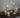 A set of white angel-shaped candle holders with built-in LED lights, displayed on a dark wooden table against a soft gray wall with delicate leafy branches.