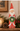 A rustic Santa Claus figurine holding a green wreath, displayed on a wooden surface with a lit candle, pinecone, and holiday greenery in the background.